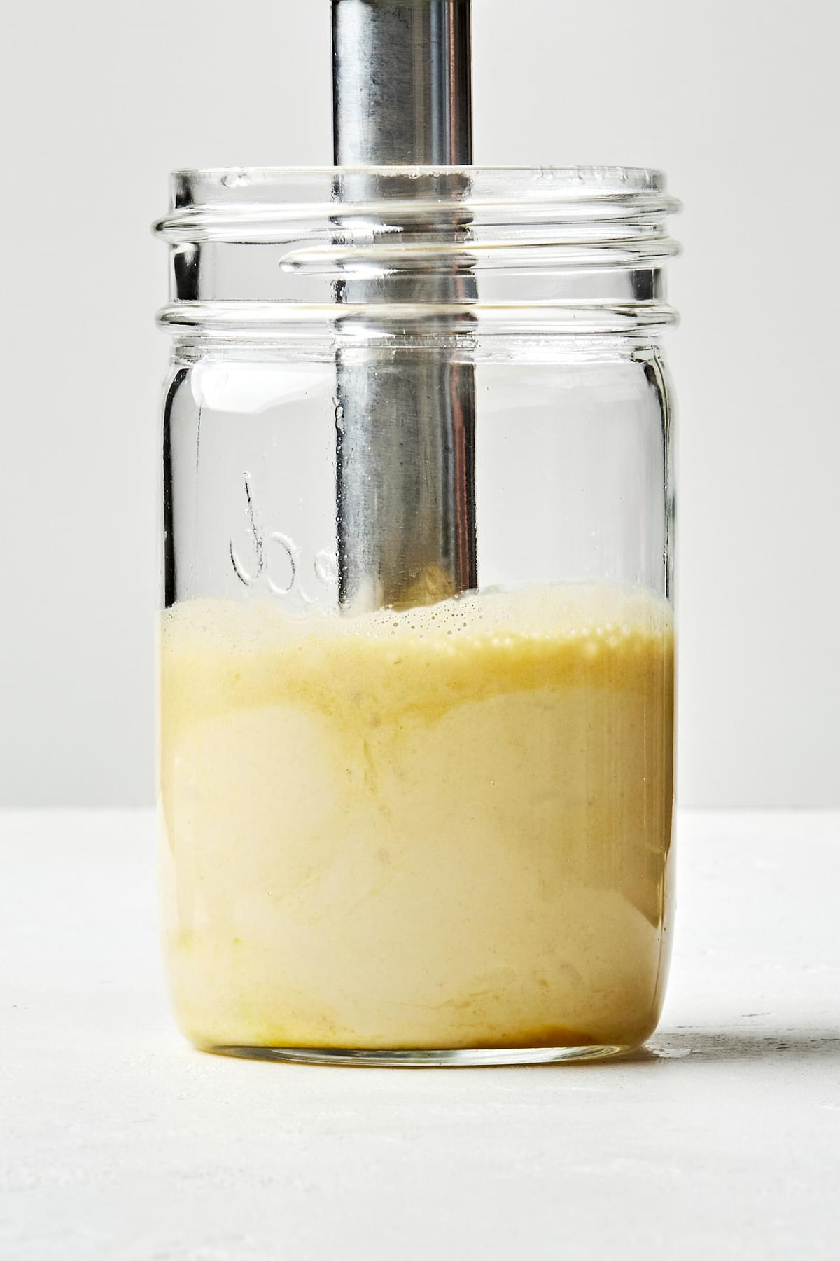 homemade caesar dressing being blended together in a glass jar with an immersion blender