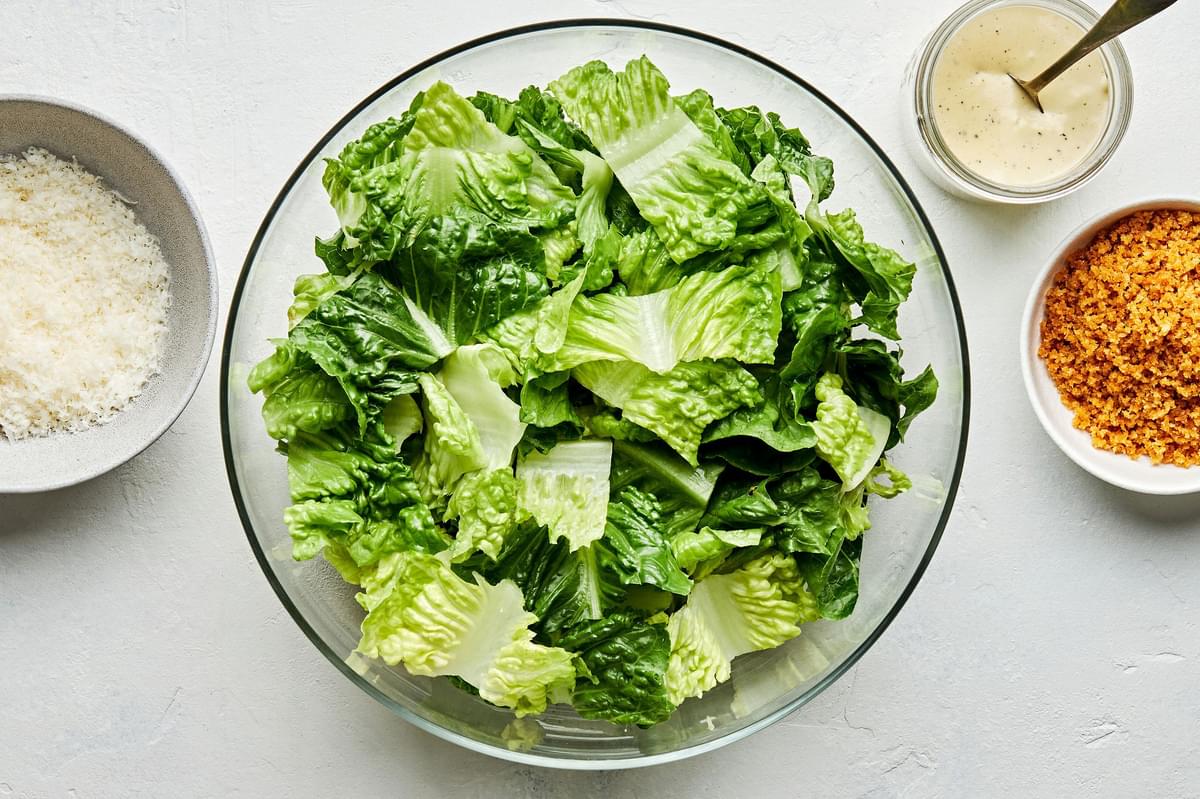 romaine lettuce in a salad bowl surrounded by bowls of grated parmesan, toasted breadcrumbs and homemade caesar dressing