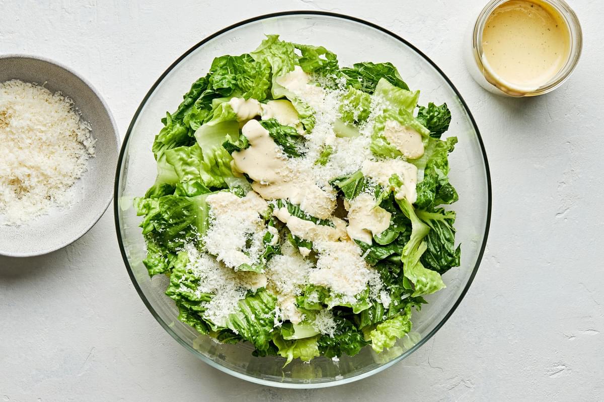 chopped romaine in a salad bowl topped with homemade caesar dressing and parmesan next to bowls extra parmesan and dressing