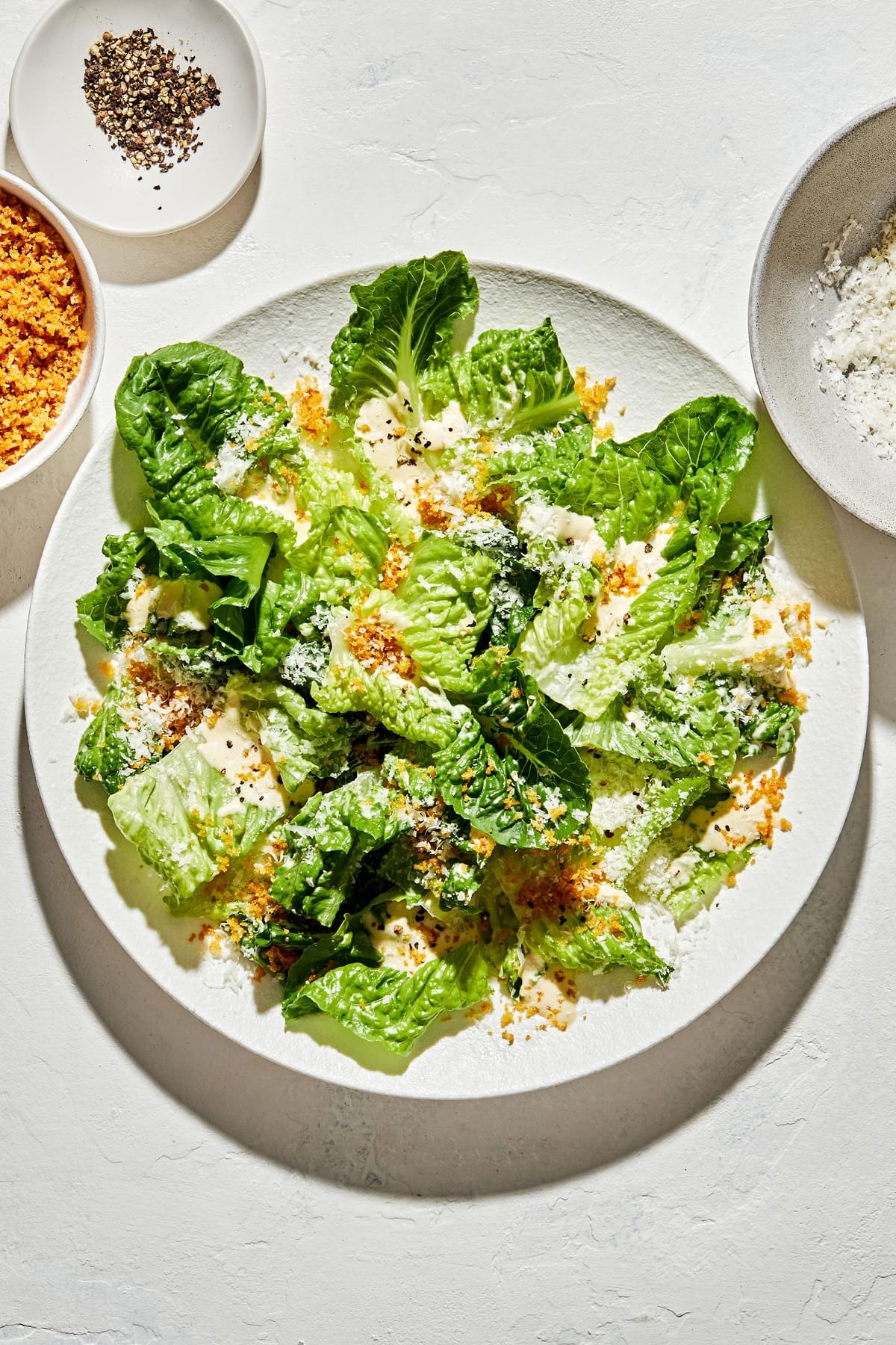 homemade Caesar salad with homemade caesar dressing, surrounded by bowls of toasted breadcrumbs, pepper and Parmesan