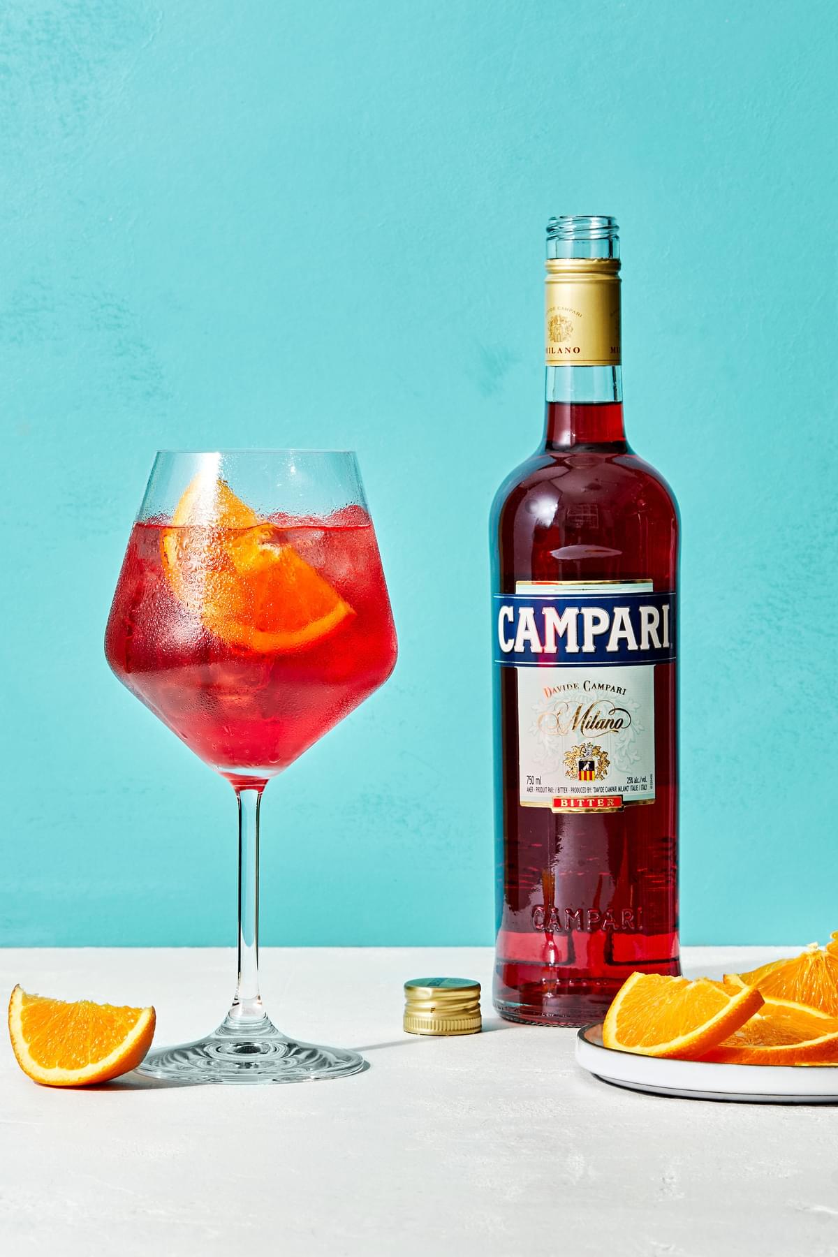 a campari spritz in a wine glass garnished with an orange wedge next to a bottle of campari and a plate of orange wedges