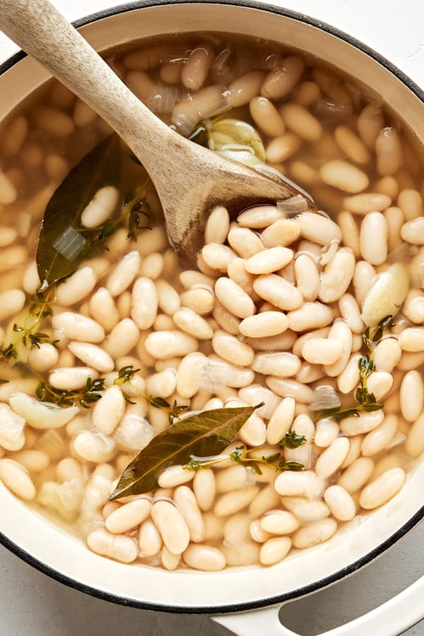 a pot of cannellini beans made with olive oil, onion, bay leaves, thyme, garlic, salt & pepper being stirred with a spoon