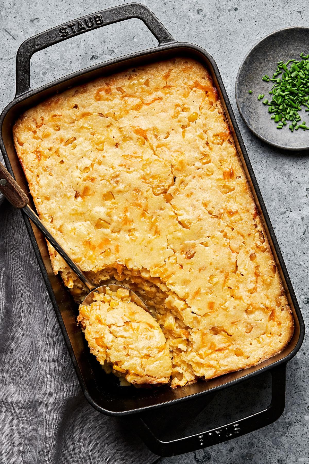 cheesy corn casserole in a casserole dish with a spoon scooping out a serving