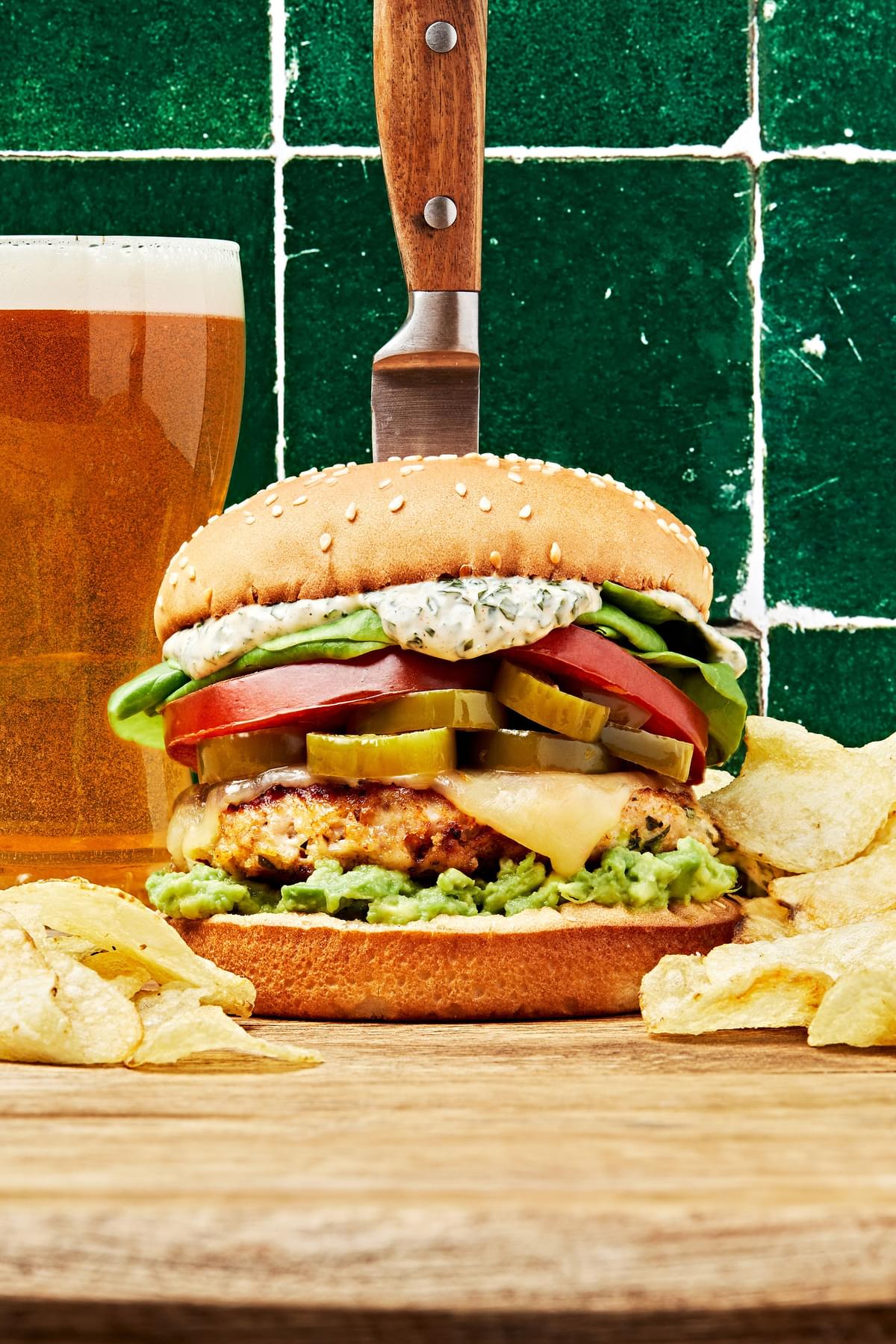 chicken burger on a bread board with a knife sticking in it surrounded by potato chips and a glass of beer
