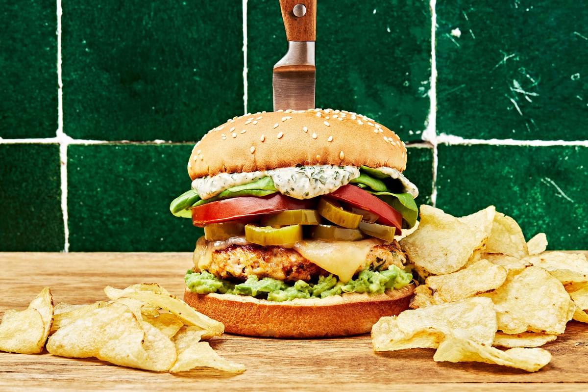 chicken burger with pepper jack, basil mayo, avocado spread and candied jalapeños with a knife in it next to potato chips