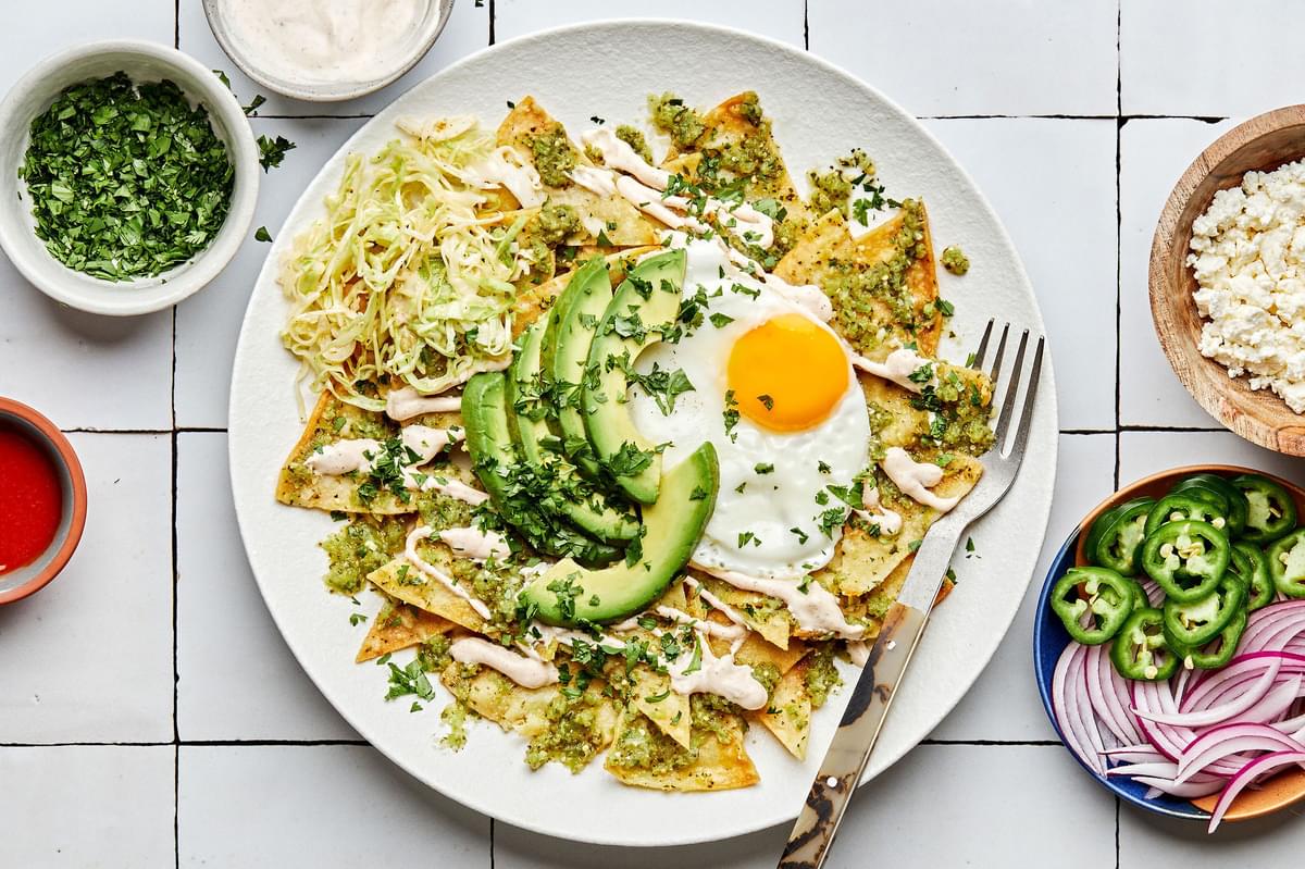 chilaquiles on a plate with a fork surrounded by bowls of toppings: cilantro, red onion, jalapeño, queso fresco and hot sauce