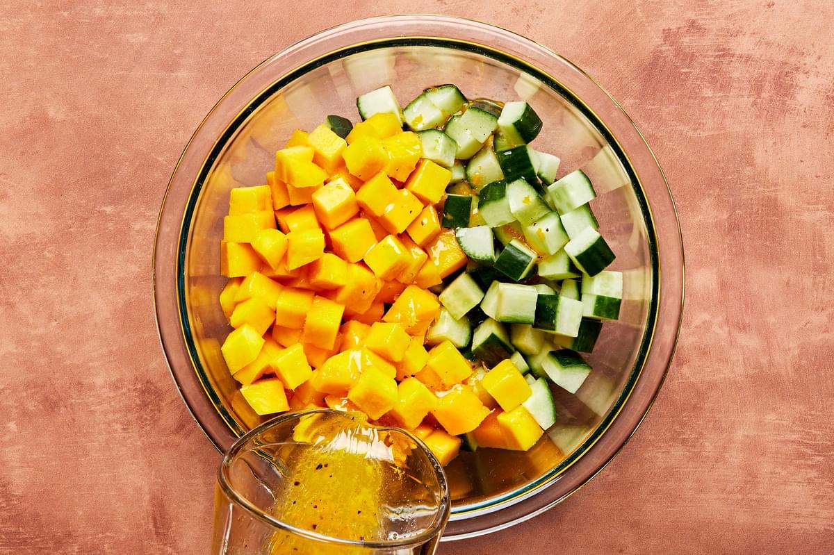 homemade citrus marinade being poured over diced English cucumbers and mangoes in a glass bowl