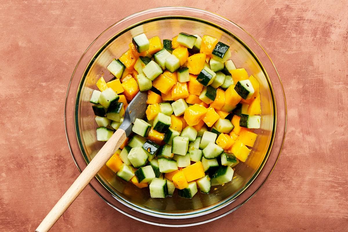 Diced English cucumbers, mangoes and homemade citrus marinade being stirred together in a glass bowl with a wooden spoon