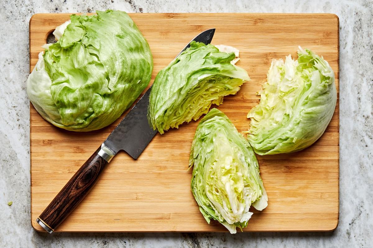 a head of iceberg lettuce being chopped into wedges with a knife on a wooden cutting board