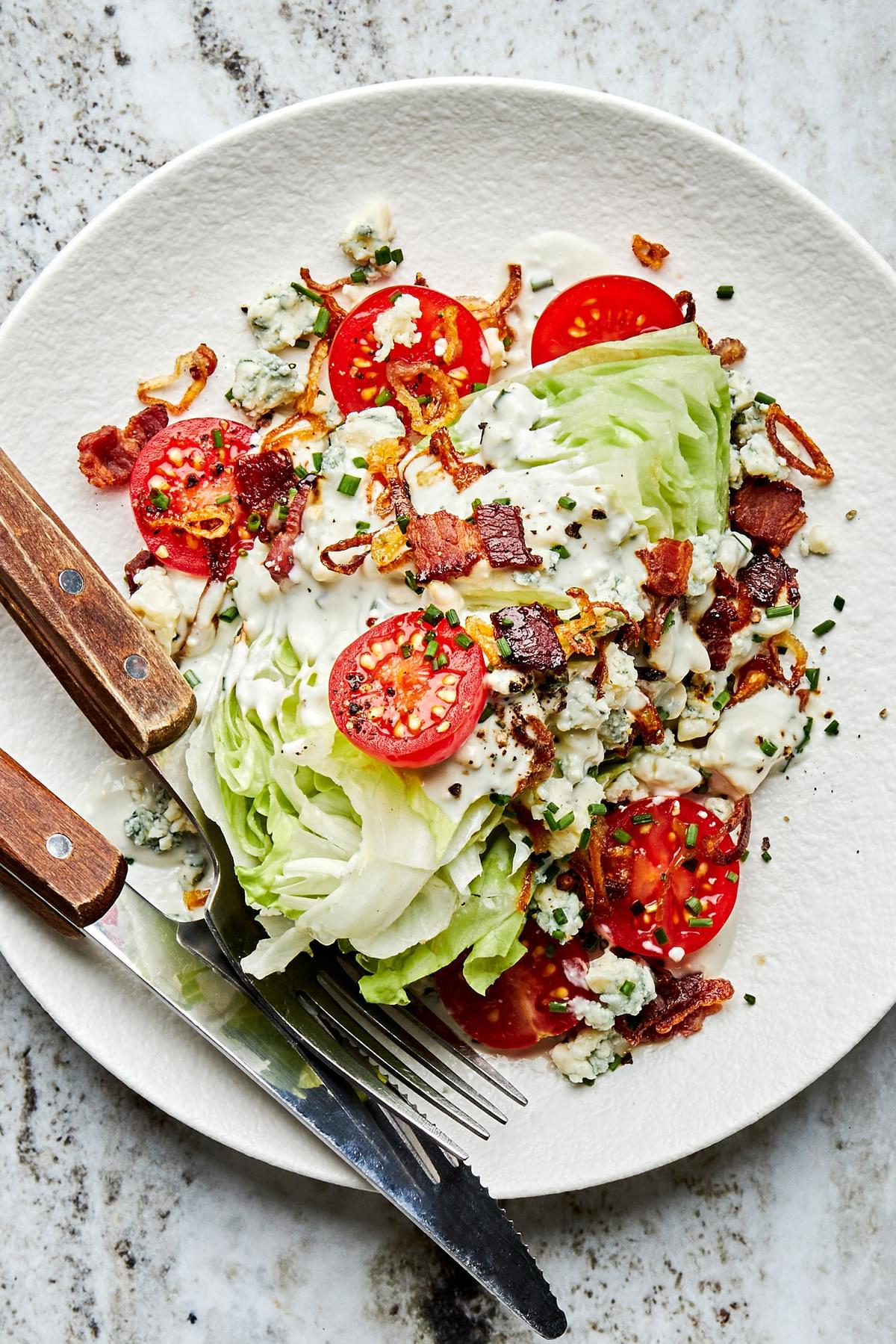a classic wedge salad with homemade blue cheese dressing, bacon, tomatoes and fried shallots on a plate with a fork and knife