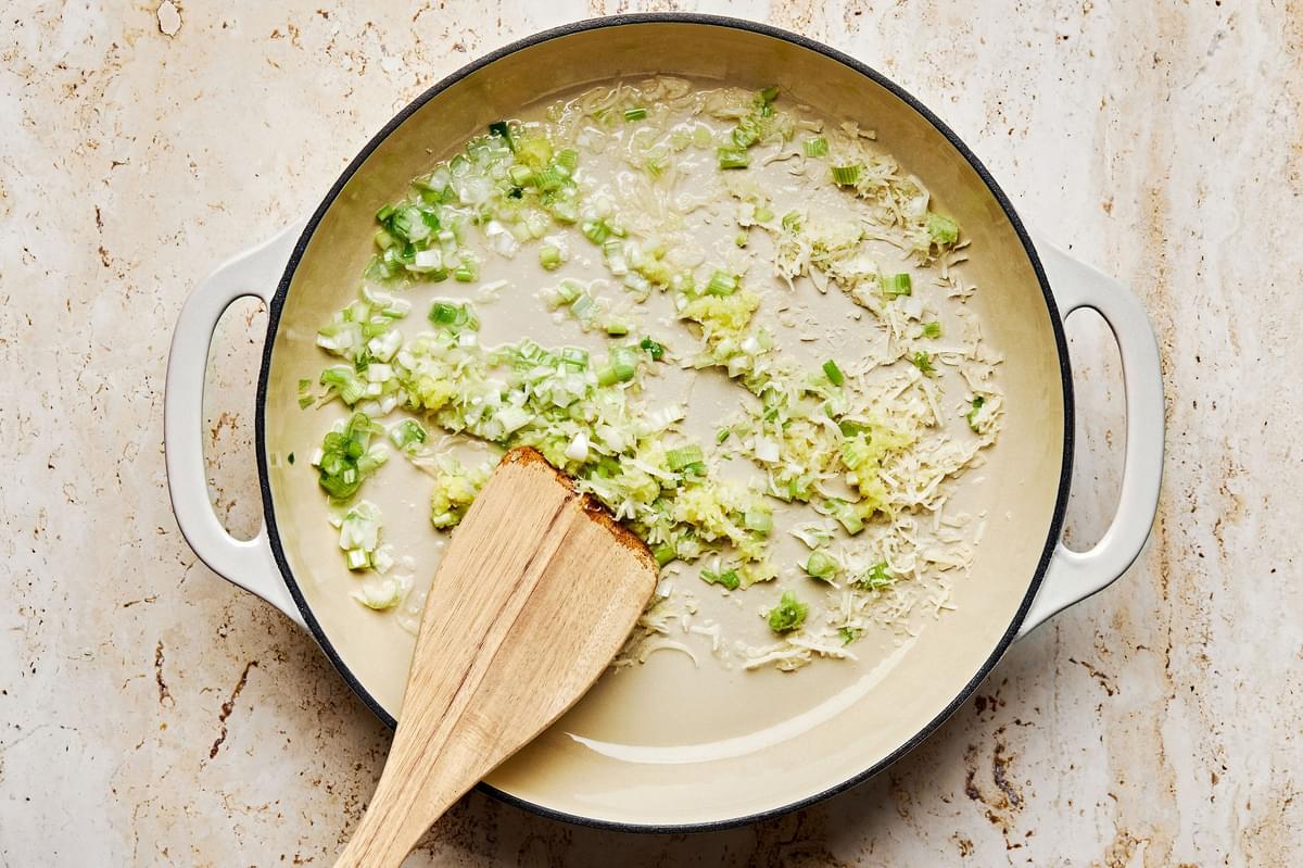 white and light green onions, coconut flakes and ginger being cooked in coconut oil in a pan with a wooden spoon