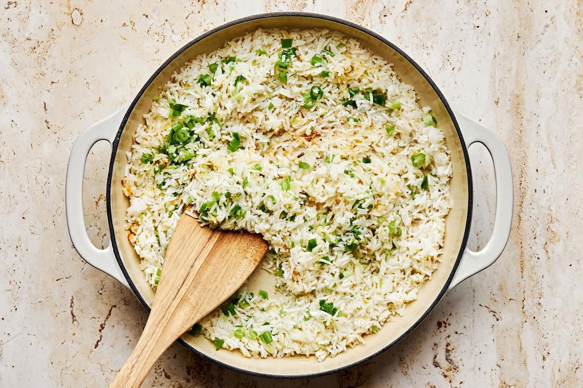 coconut rice, green onions, coconut flakes and ginger being cooked in coconut oil in a pan with a wooden spoon
