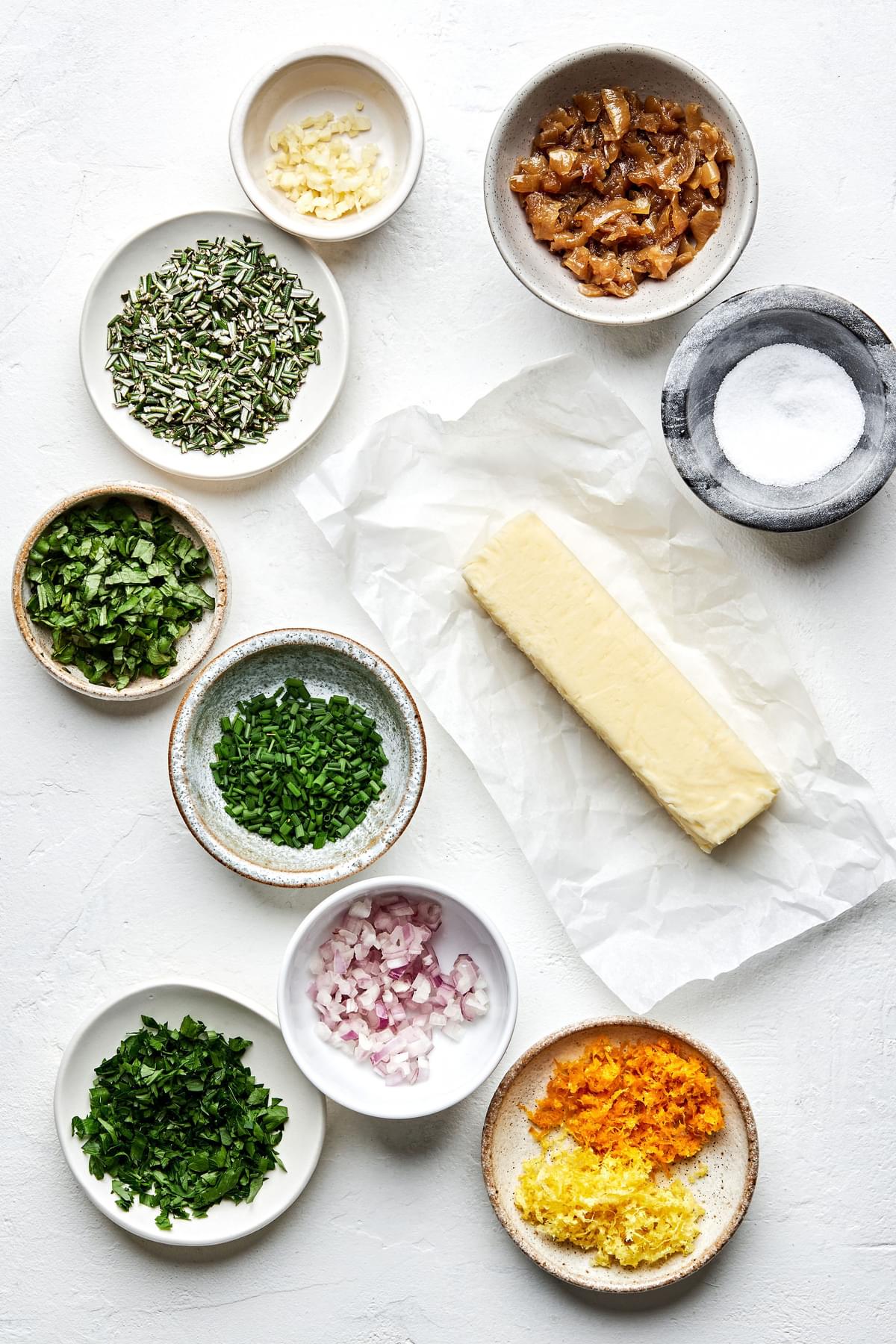 a stick of butter surrounded by bowls of salt, caramelized onion, citrus zest and fresh herbs for making compound butters