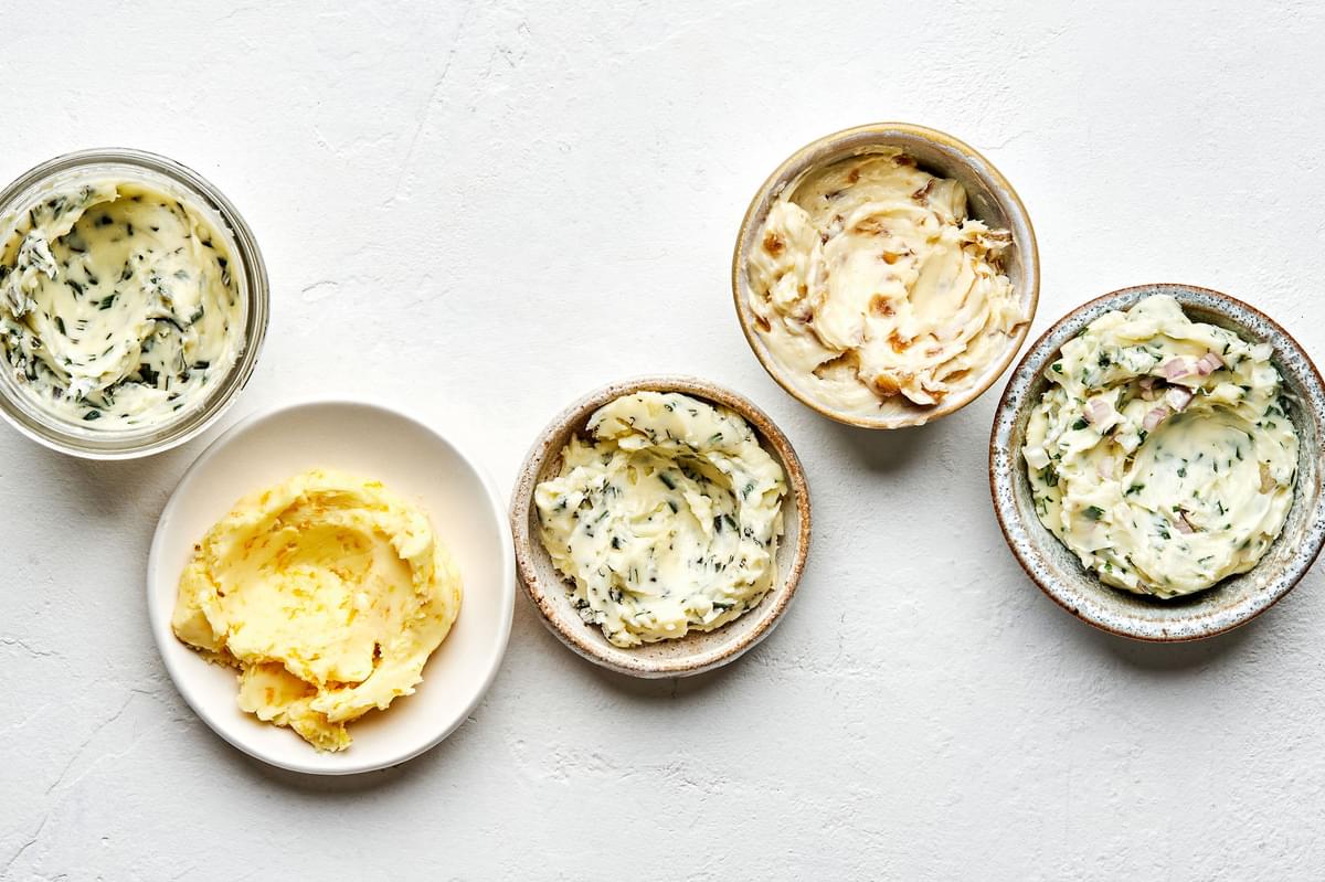 5 bowls of softened homemade compound butters made with fresh herbs, citrus zest and juice and caramelized onions