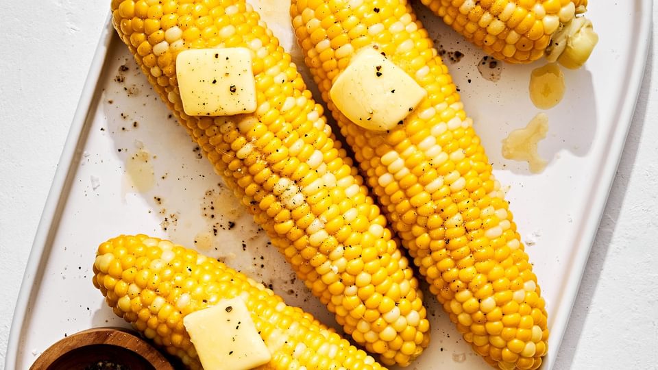 4 homemade corn on the cob on a platter topped with butter beside small bowls of salt and pepper for serving