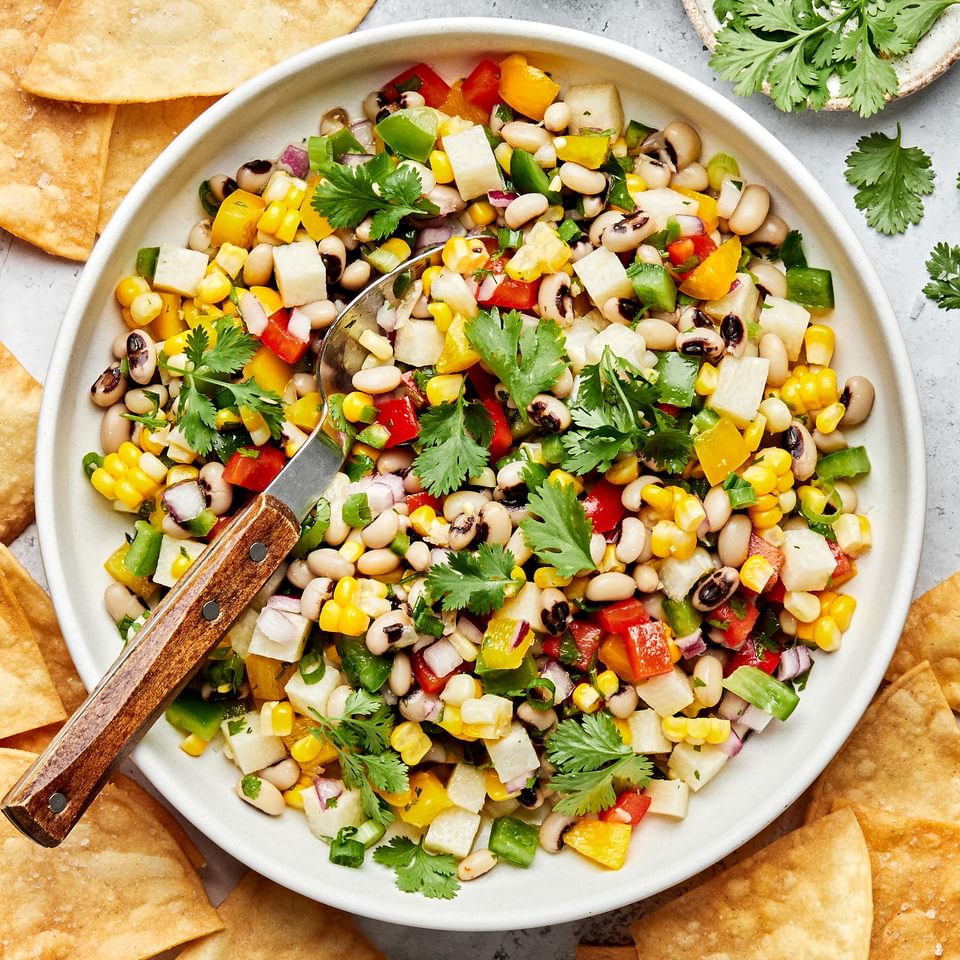 homemade cowboy caviar in a serving bowl with a spoon surrounded by tortilla chips and bowls of salt and cilantro