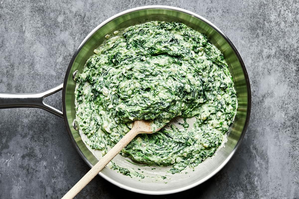 creamed spinach being stirred in a skillet with a wooden spoon made with butter, cream, dijon, spices and parmesan cheese