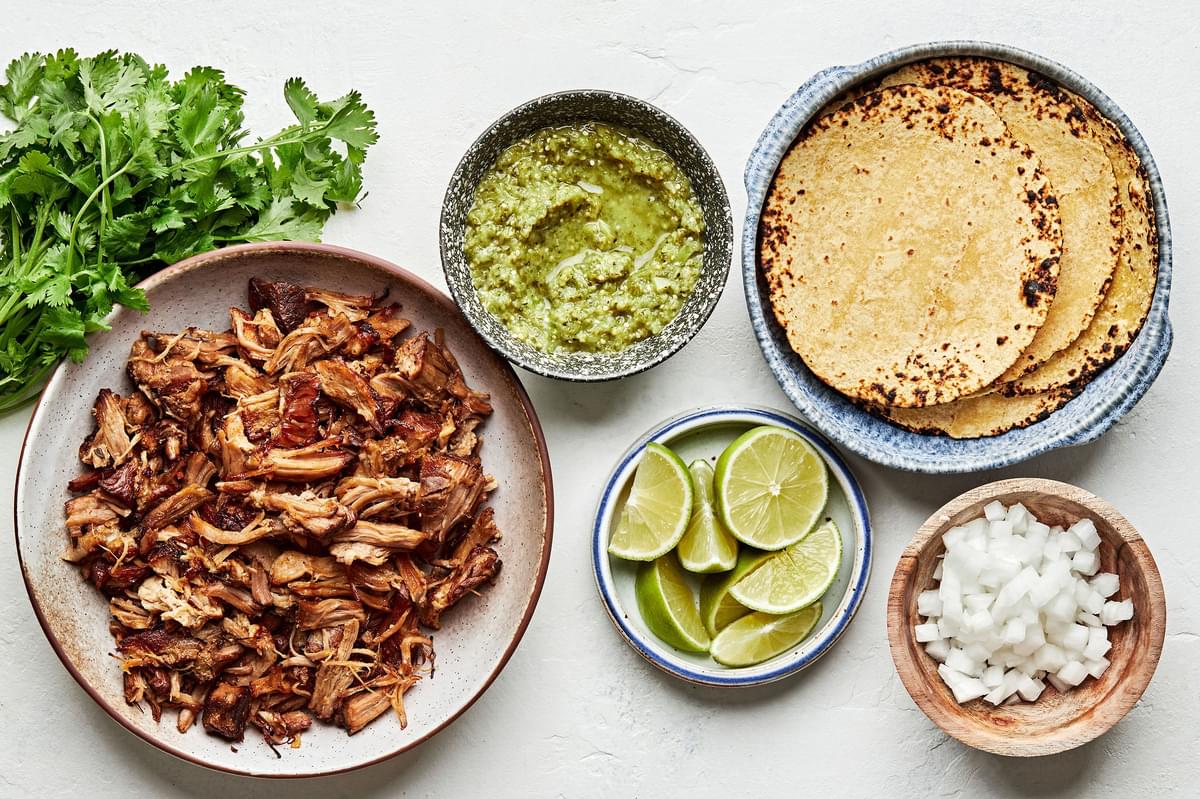 crispy pork carnitas, corn tortillas, salsa verde, onion, cilantro and lime wedges in bowls to make crispy carnitas tacos