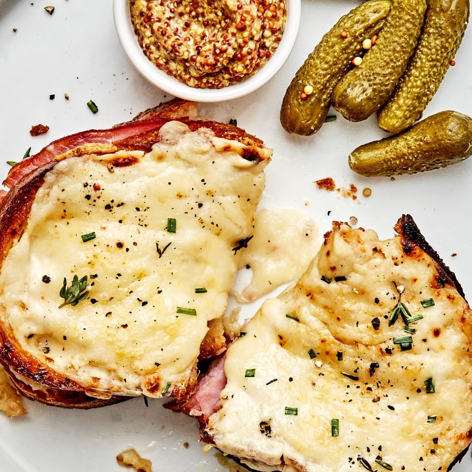a homemade croque monsieur sandwich on a plate served with pickles and stone ground mustard