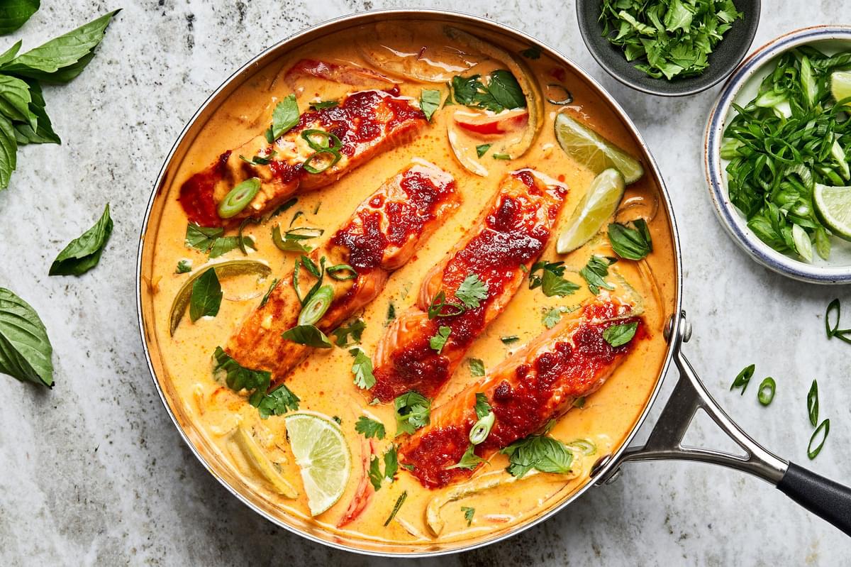 Coconut curry in a large skillet with four filets of salmon topped with cilantro and basil and fresh lime wedges.