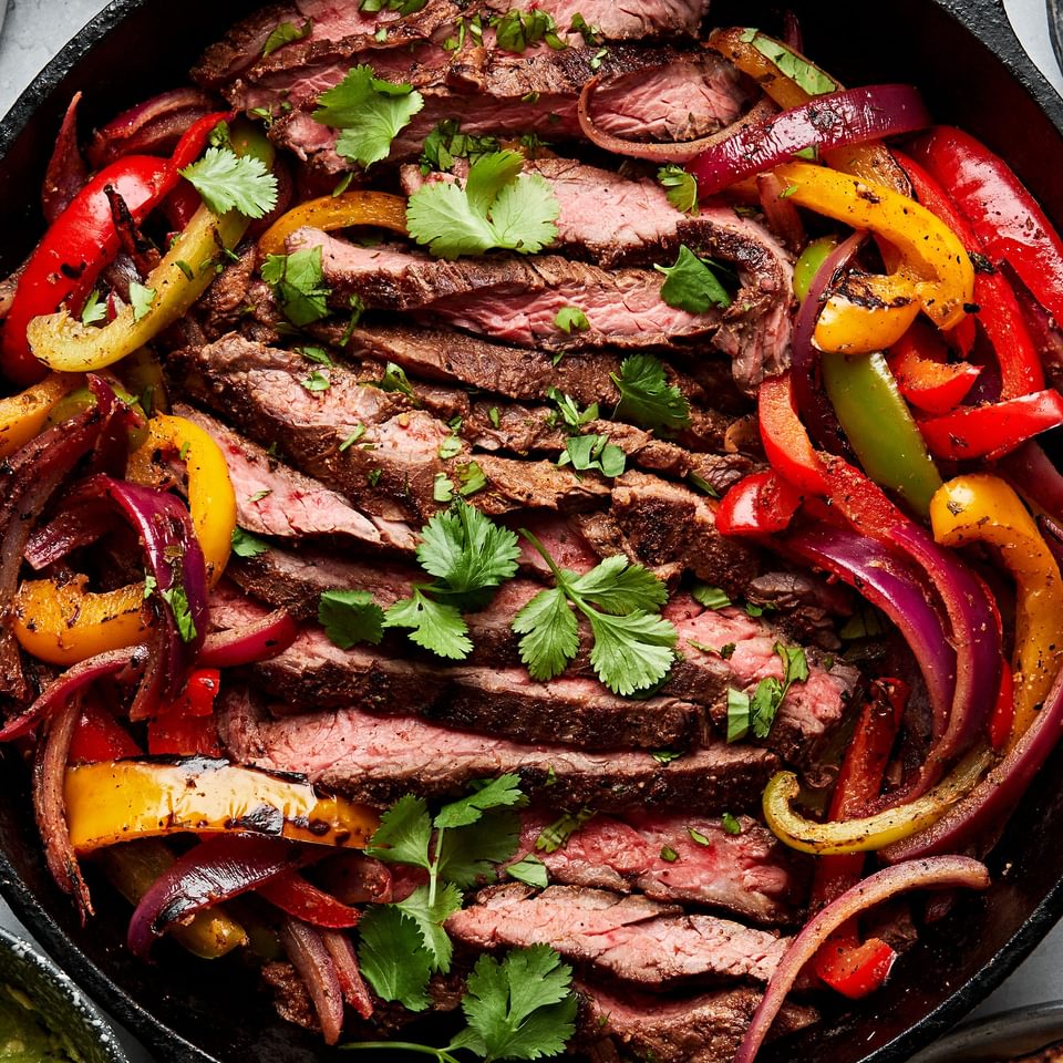 steak fajitas and peppers in a cast iron skillet sprinkled with cilantro surrounded by salsa, guacamole and corn tortillas
