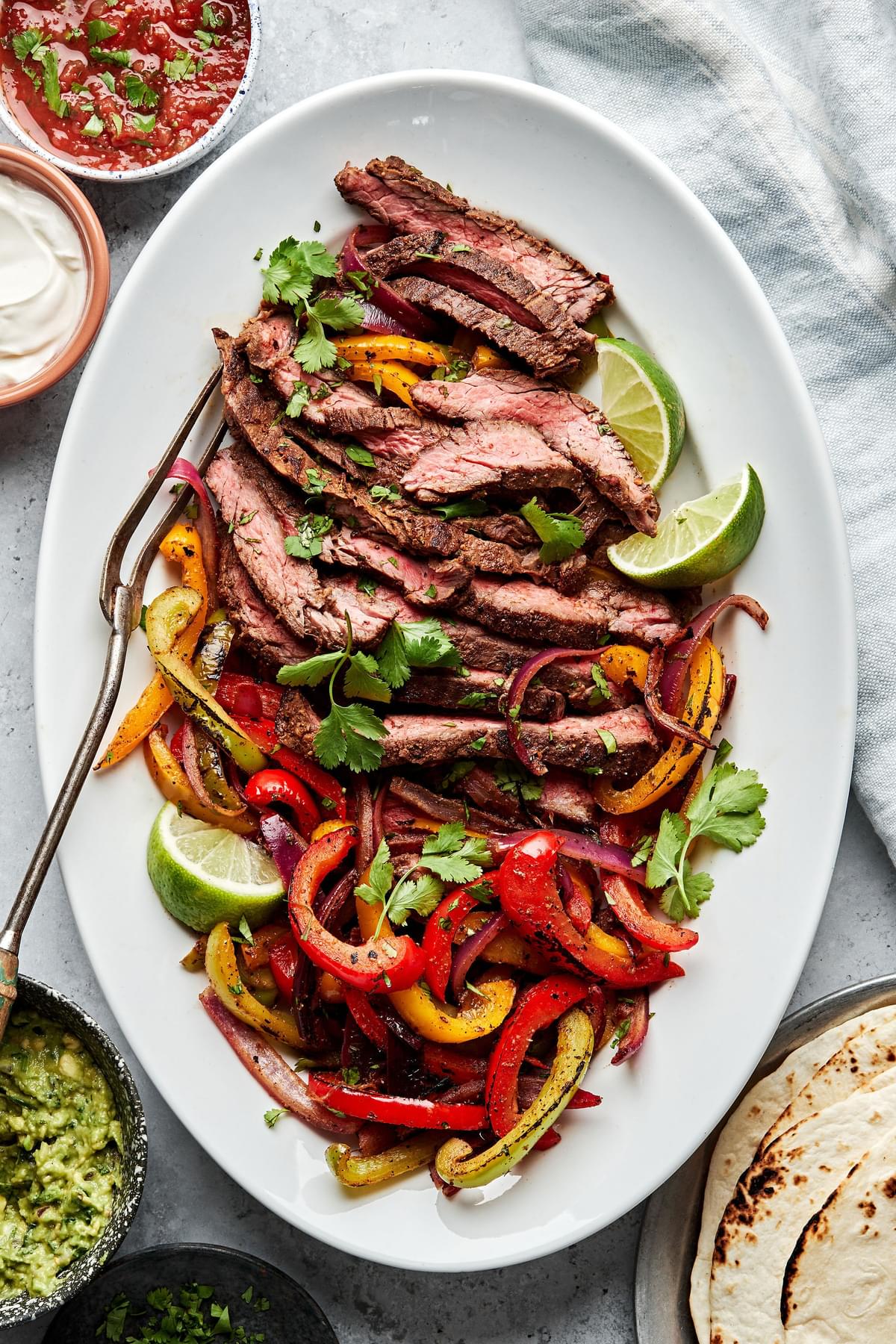 steak fajitas on a serving plate sprinkled with cilantro surrounded by salsa, guacamole, sour cream and corn tortillas