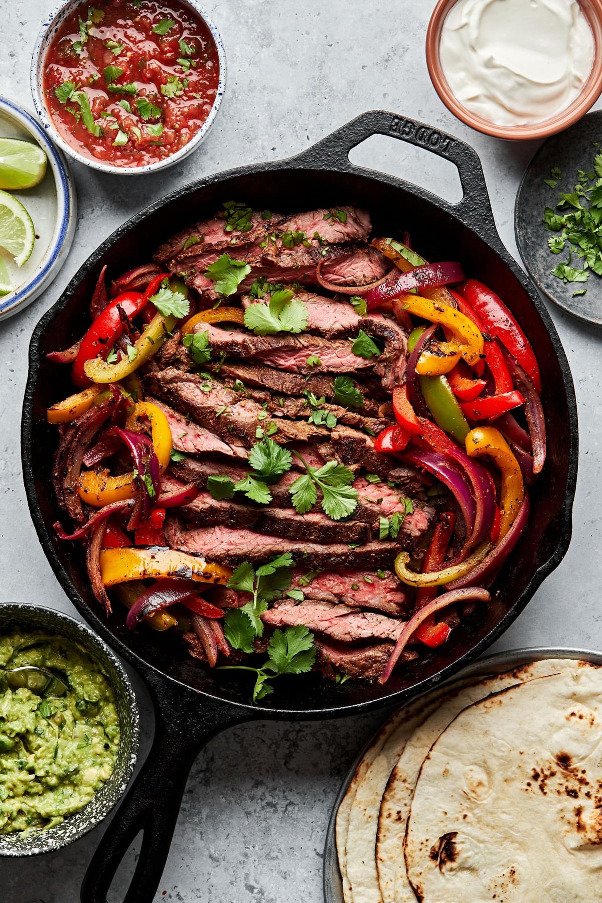 steak fajitas and bell peppers in cast iron skillet sprinkled with cilantro surrounded by salsa, guacamole and corn tortillas