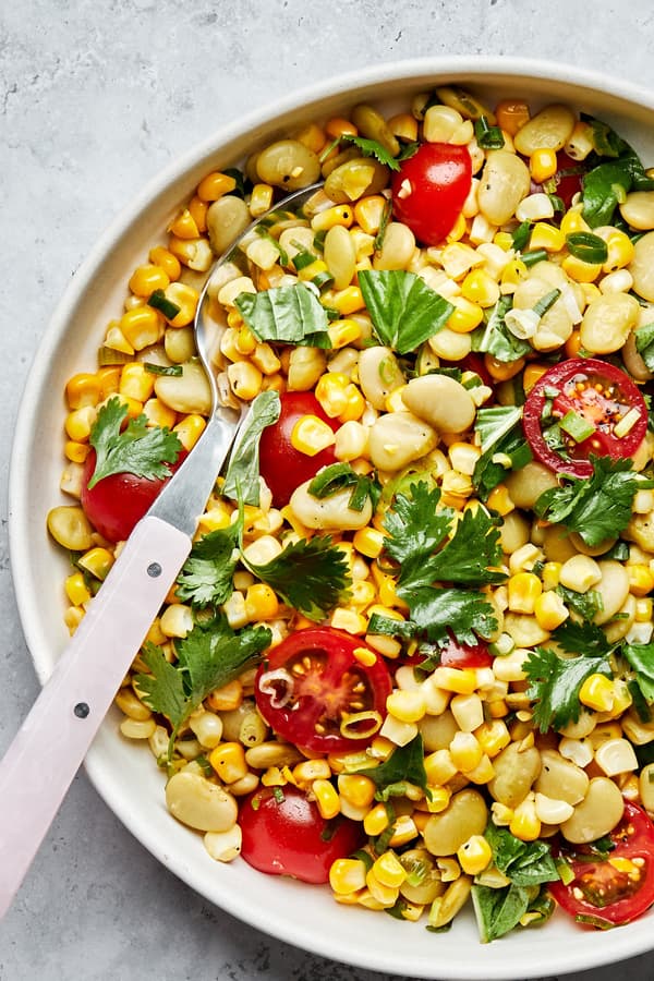 easy homemade succotash in a serving bowl with a spoon made with corn, lima beans, tomatoes, green onions, cilantro and basil