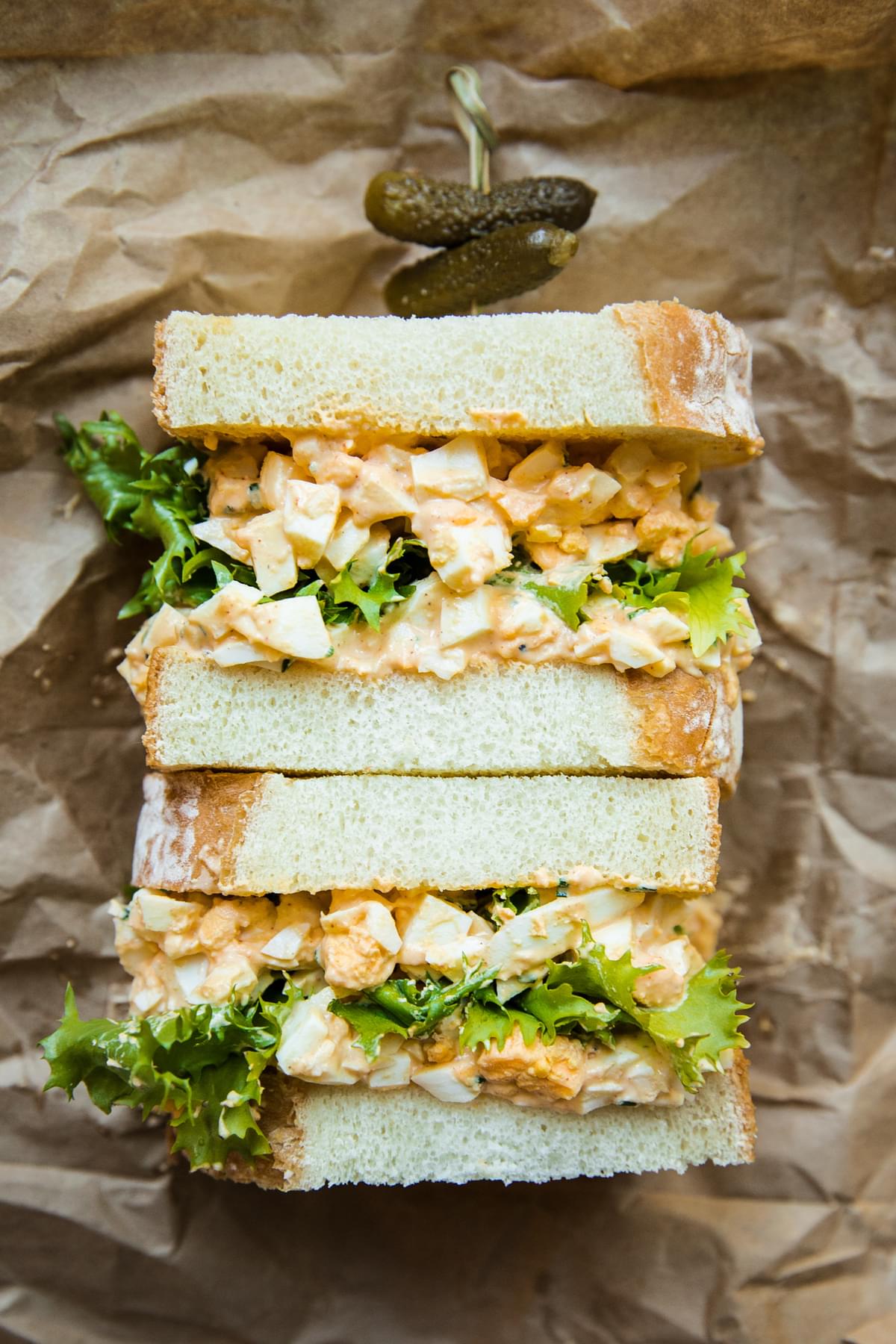 an egg salad sandwich with romaine lettuce cut in half on a piece of parchment paper with small pickles on a toothpick on top