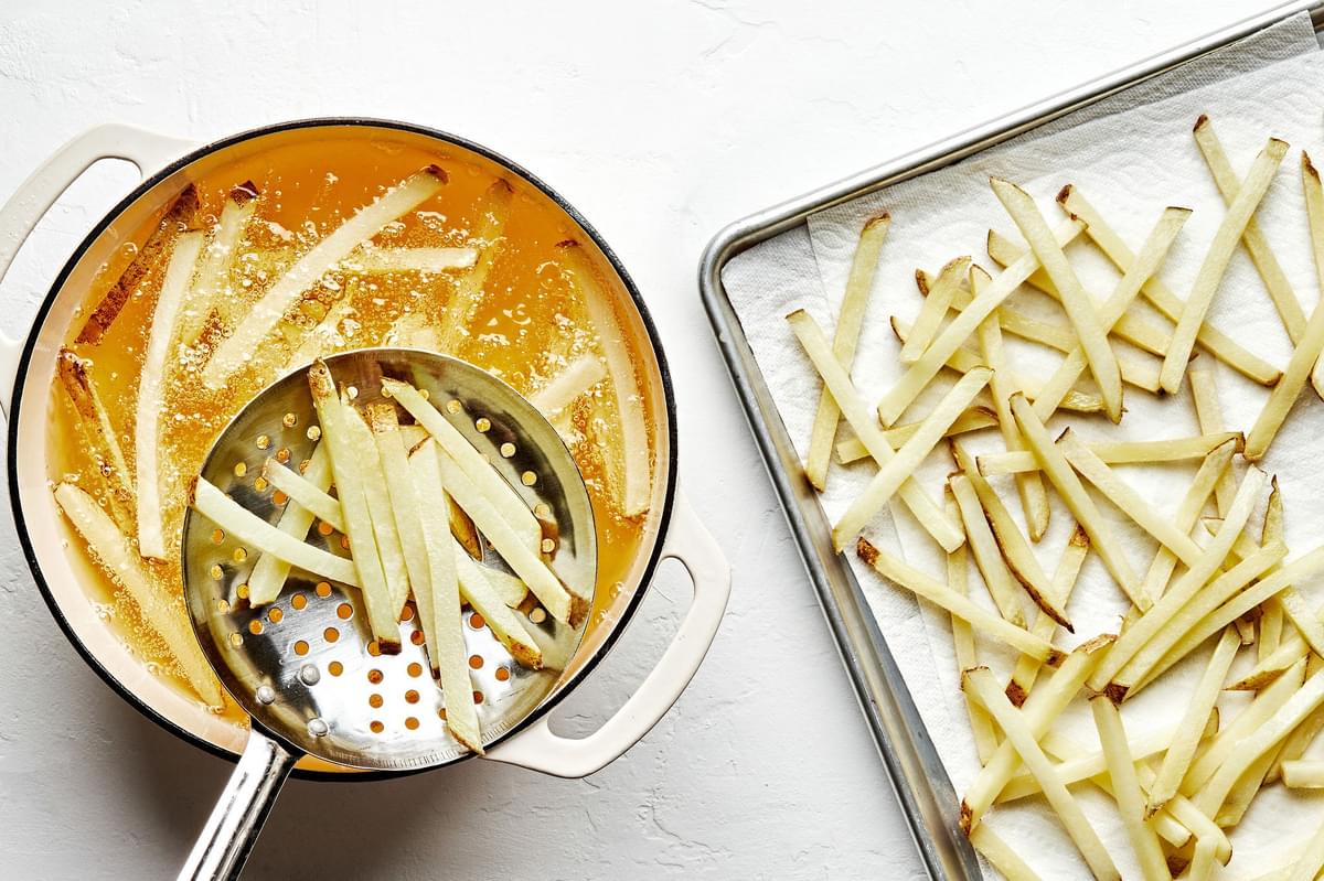 homemade fries being fried in a pot of vegetable oil and scooped onto a paper lined baking sheet to cool