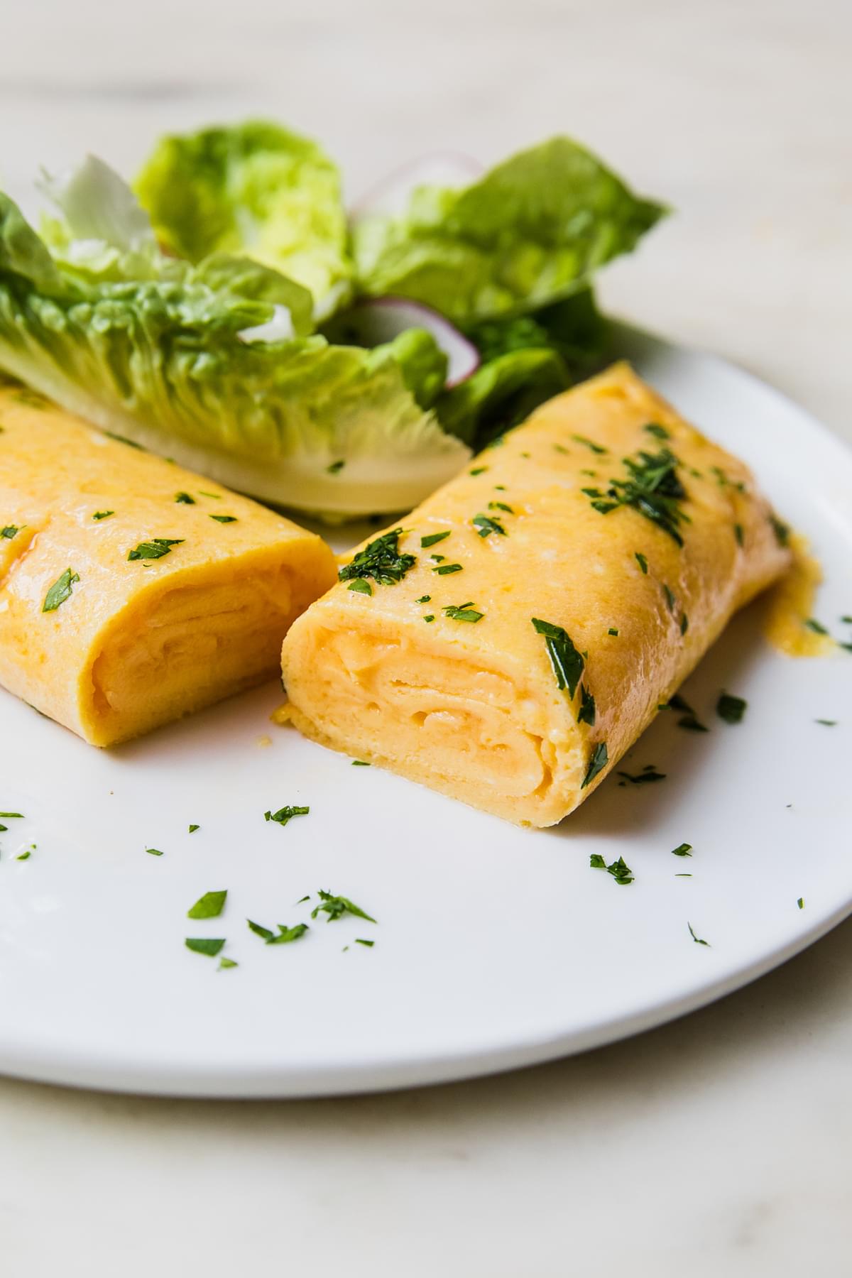 a homemade French omelette cut in half sprinkled with parsley on a plate next to a simple salad of lettuce and sliced radish