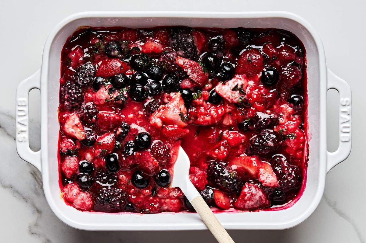 mixed fresh berries and mint leaves that have been baked in the oven being stirred with a spatula in a 9x13 pan