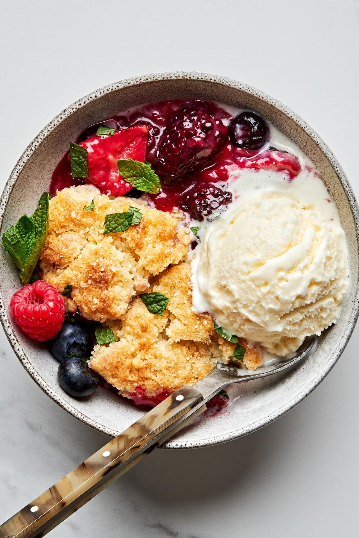 a piece of homemade fresh berry cobbler topped with a scoop of vanilla ice cream  and mint leaves in a bowl with a spoon