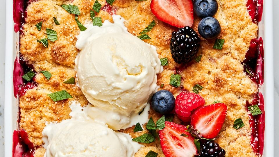 fresh berry cobbler in a 9x13 baking dish topped with 2 scoops of vanilla ice cream, additional fresh berries and fresh mint