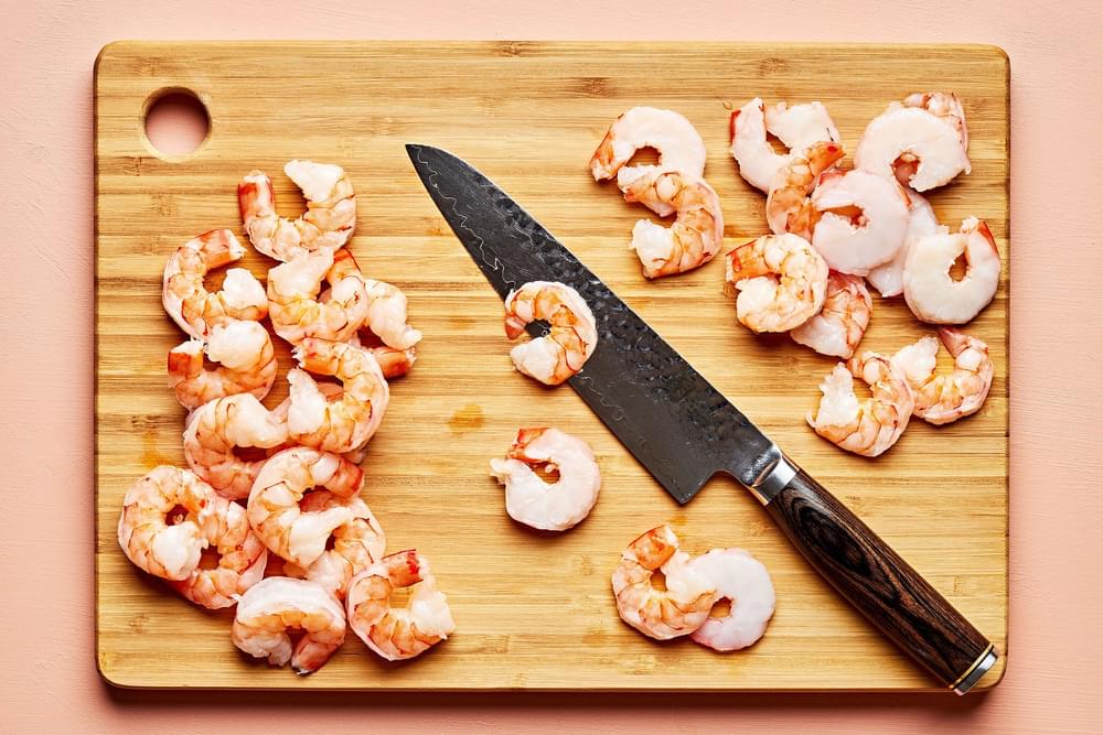 cooked shrimp being sliced crosswise  with a knife on a cutting board