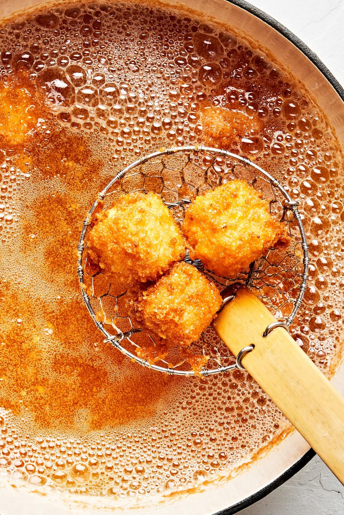 Fried cheese breaded with flour, spices, eggs and panko being scooped out of a pot of vegetable with a spider strainer