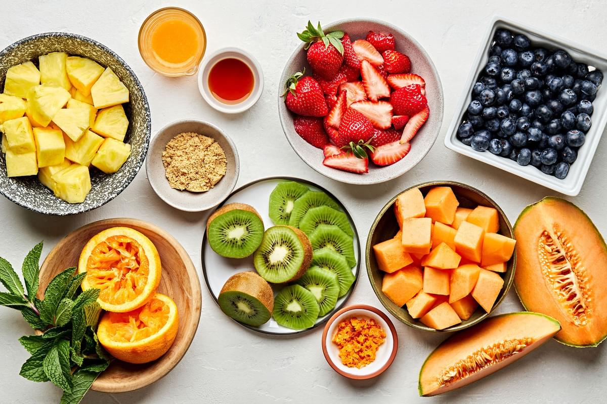Cantaloupe, Pineapple , Strawberries, Blueberries, Kiwis, Brown sugar, Mint, Orange & Vanilla in bowls for making fruit salad