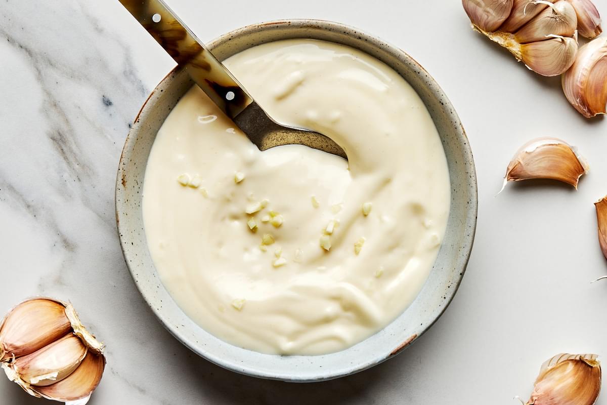 homemade garlic aioli in a bowl with a spoon surrounded by garlic cloves on the counter