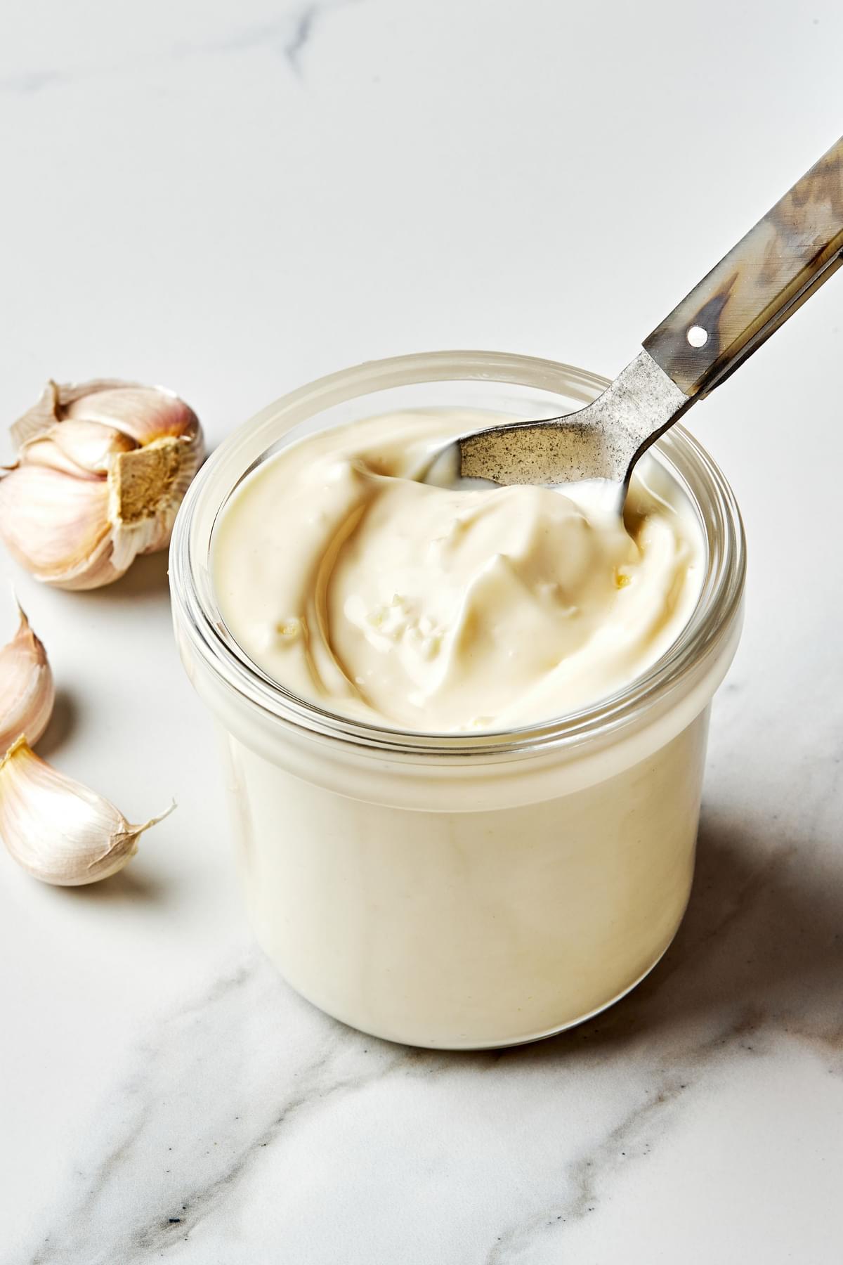 homemade garlic aioli in a jar with a spoon surrounded by garlic cloves scattered on the counter