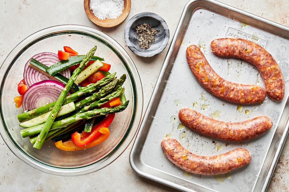 sausage on a baking sheet and raw veggies in a bowl next to bowls of salt and pepper to make grilled sausage and vegetables