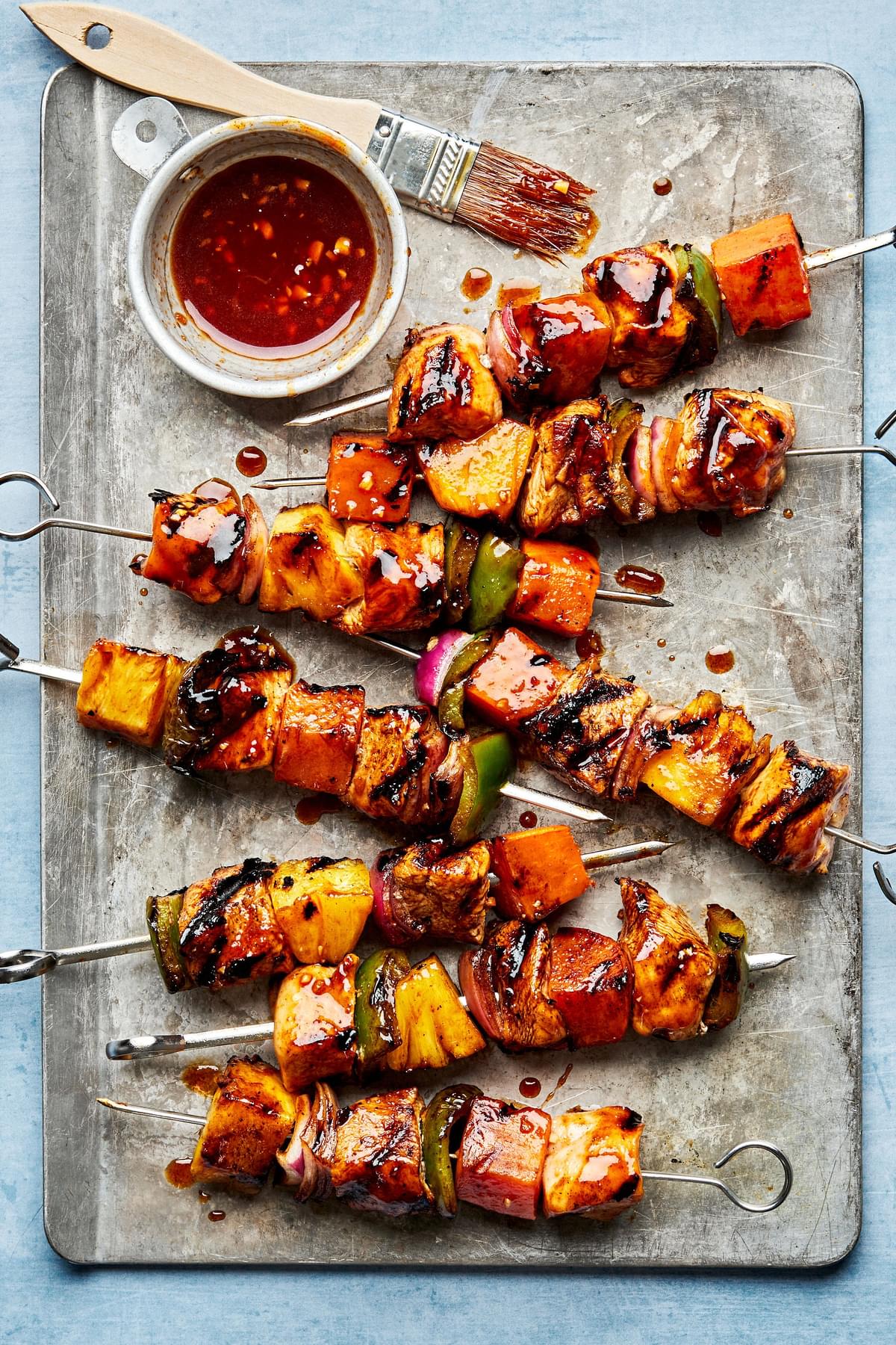 grilled Hawaiian chicken kabobs on a baking sheet brushed with remaining marinade next to a bowl of additional marinade