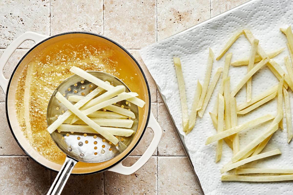 homemade French fries being deep fried fried in a pot of peanut oil and being removed with a slotted spoon onto a paper towel