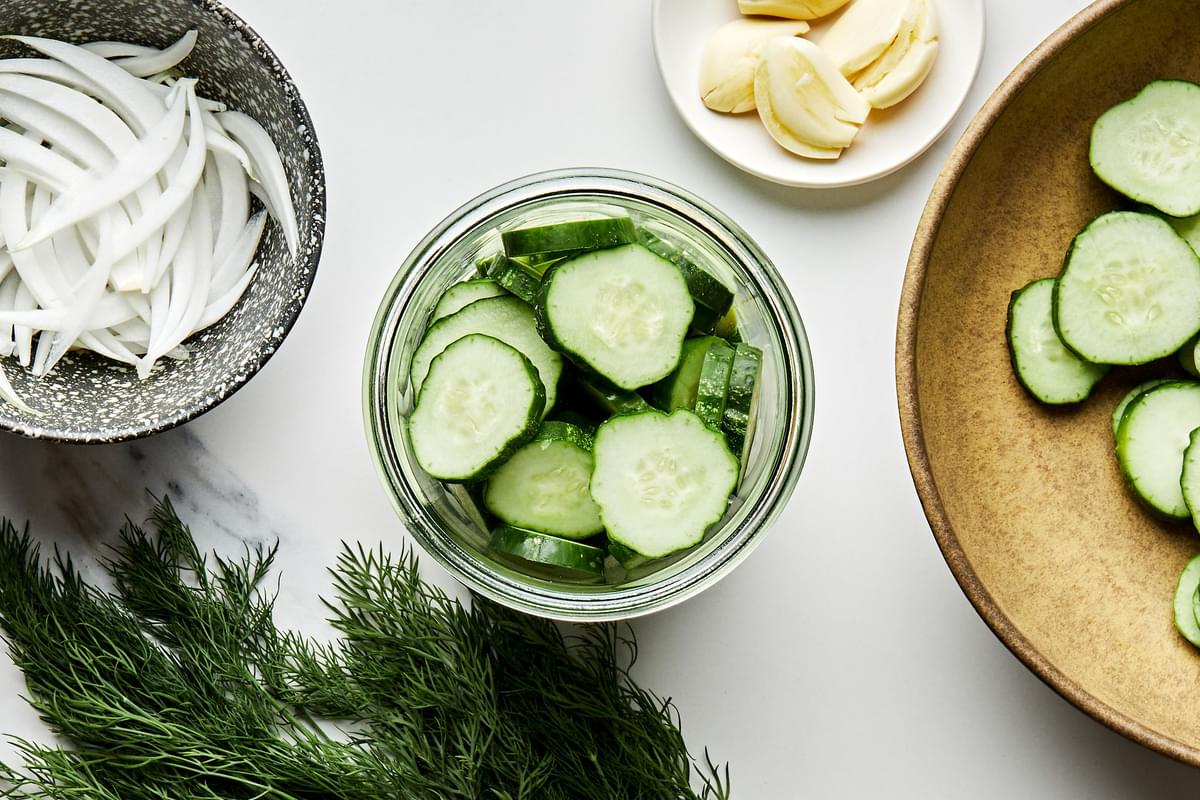 sliced sweet onion and sliced cucumbers in a jar surrounded by bowls of sliced onions, cucumbers, garlic and dill