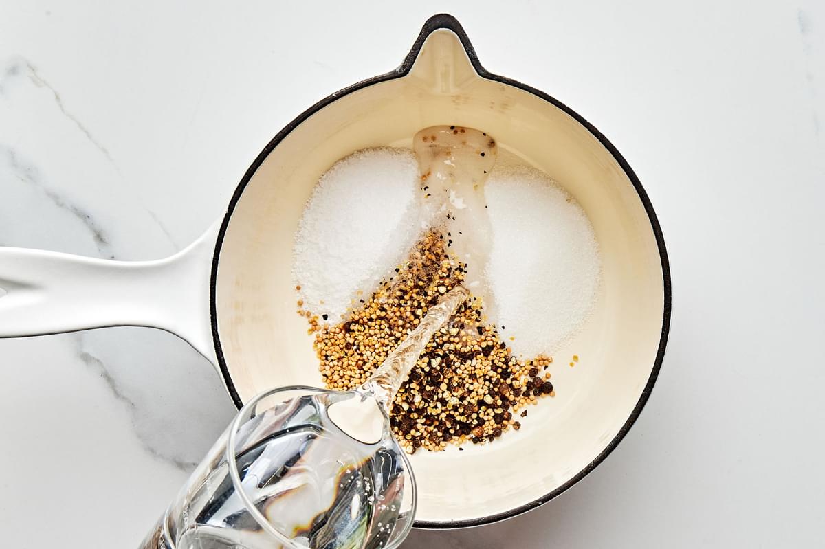 water being poured into a sauce pan with crushed mustard seeds, peppercorns, vinegar, sugar and salt