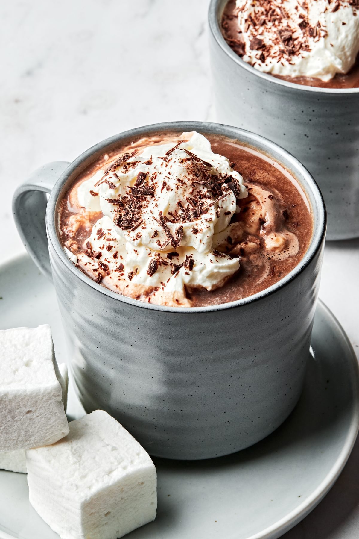2 mugs of homemade hot chocolate topped with whipped cream and chocolate chips served with marshmallows