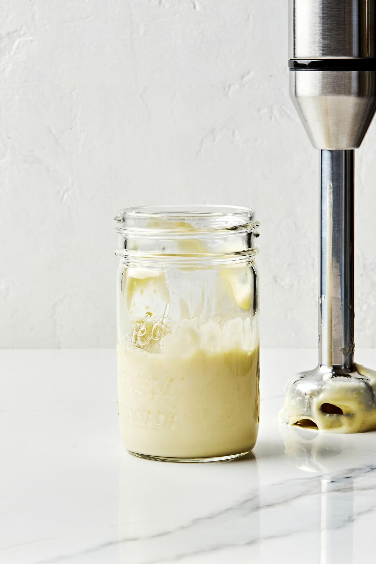 a glass mason jar of homemade mayonnaise sitting next to an immersion blender covered in homemade mayonnaise