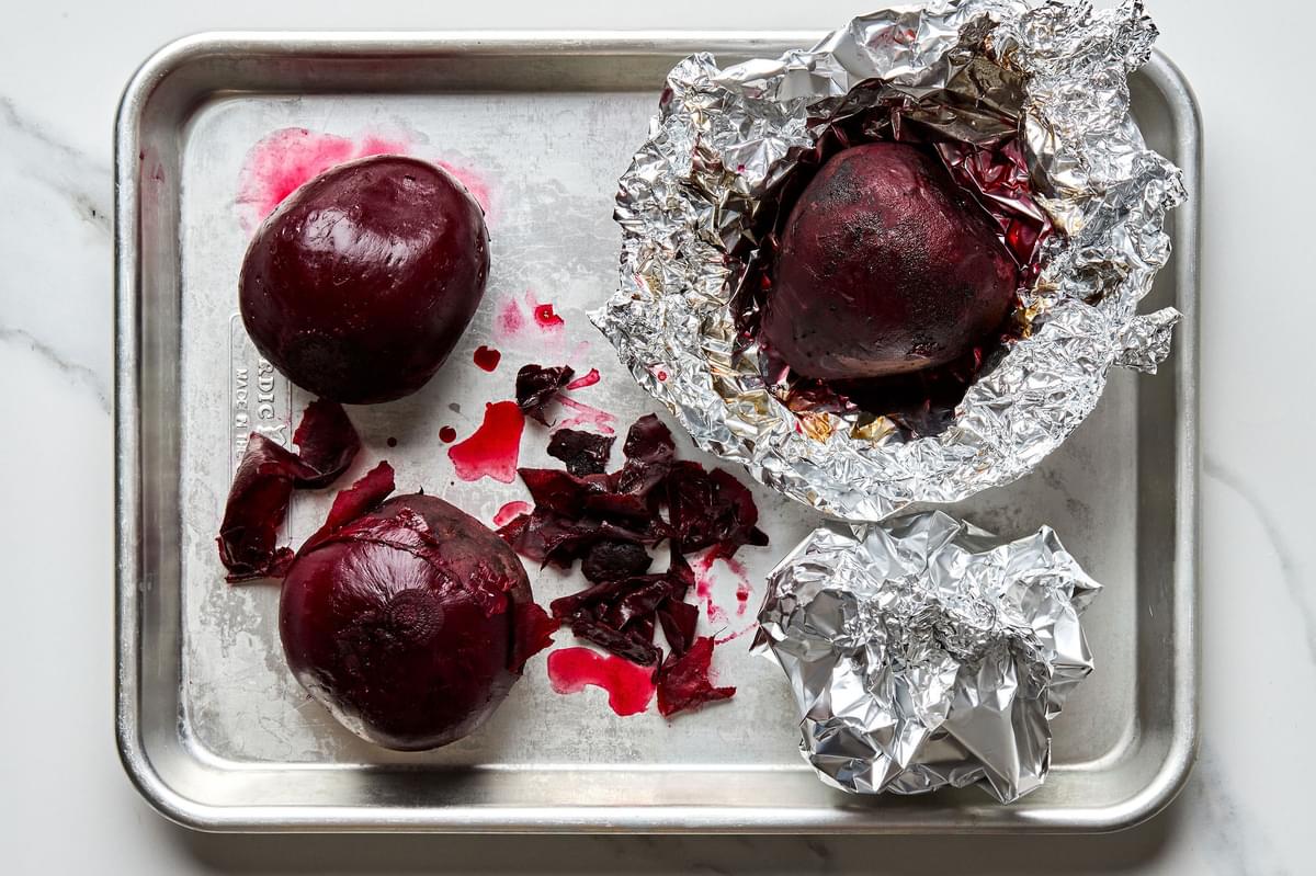 roasted beets on a baking sheet some still wrapped in foil some out of the foil with the skins being removed