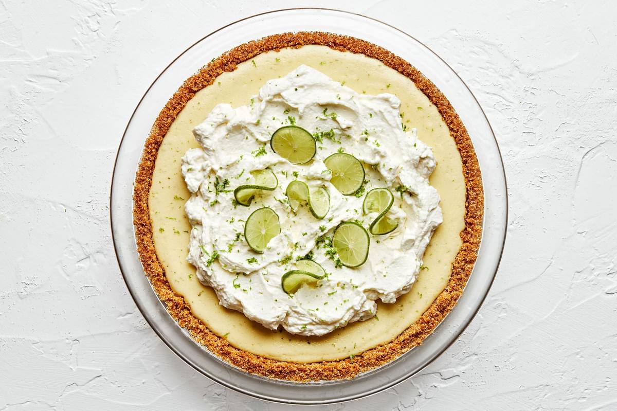 a homemade key lime pie in a glass pie pan topped with whip cream and sliced key limes