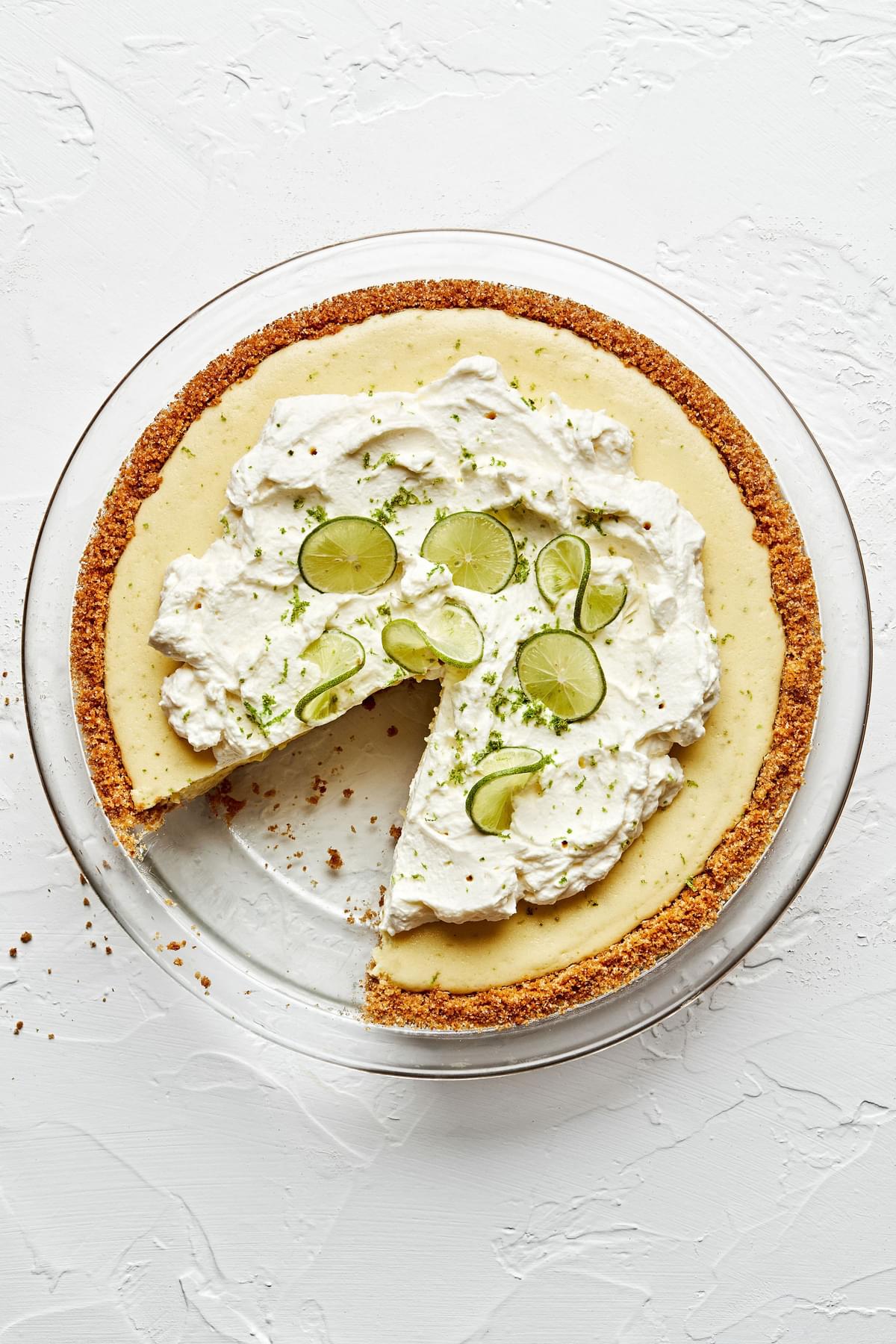 a homemade key lime pie in a glass pie pan topped with whip cream and sliced key limes with a slice cut out