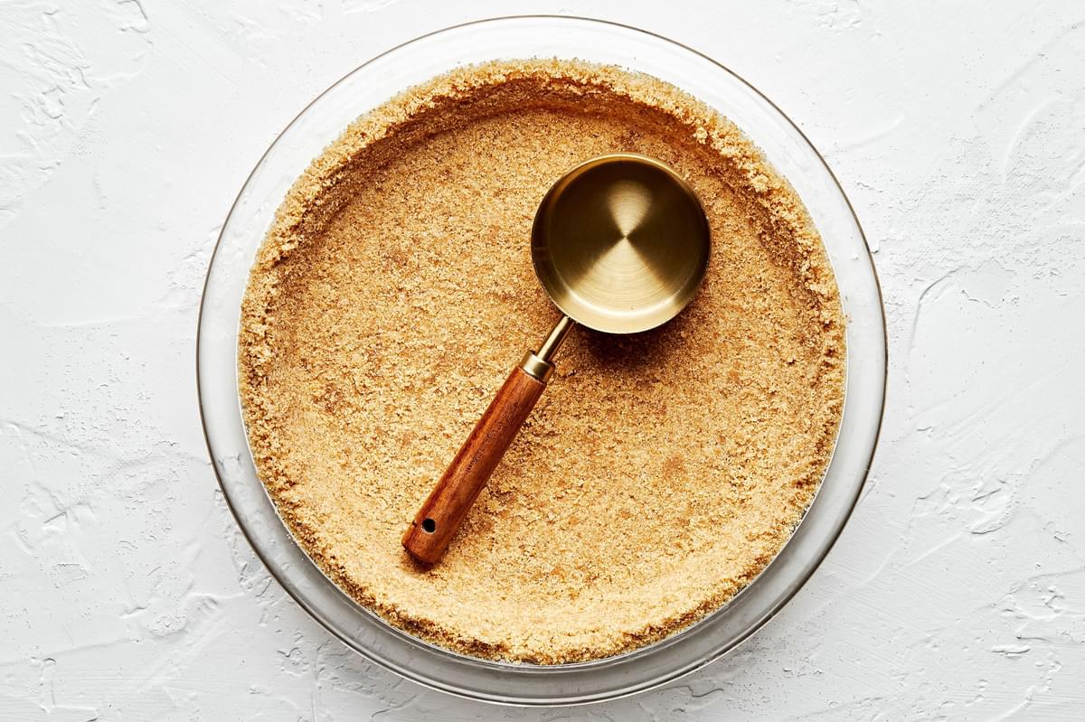 raw homemade graham cracker crust in a glass pie pan being pressed down with a measuring cup